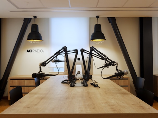 A modern radio studio setup with a long wooden table and multiple microphones mounted on adjustable arms. Two large pendant lights hang overhead, providing focused lighting. The walls are a neutral color, creating a clean and professional atmosphere. The letters 'ACI RADIO' are visible on the wall, indicating the studio's branding.