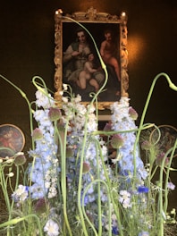 A classic painting in an ornate, golden frame is partially obscured by tall, delicately arranged white and purple flowers with lush green stems. The scene is elegantly set against a dark, textured background that highlights the vividness of the flowers and the art piece. The painting seems to depict a family group, with soft lighting that creates an intimate and serene atmosphere.