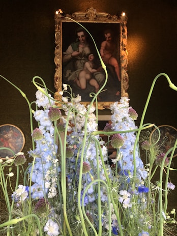 A classic painting in an ornate, golden frame is partially obscured by tall, delicately arranged white and purple flowers with lush green stems. The scene is elegantly set against a dark, textured background that highlights the vividness of the flowers and the art piece. The painting seems to depict a family group, with soft lighting that creates an intimate and serene atmosphere.