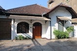 A single-story house with a tiled roof and a cream-colored facade, featuring a central wooden door with dark trim. The architecture includes arched windows and decorative gables, with potted plants lining the front. The surrounding area is paved, and there is a garage door to the left.