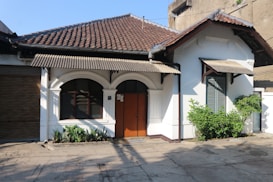 A single-story house with a tiled roof and a cream-colored facade, featuring a central wooden door with dark trim. The architecture includes arched windows and decorative gables, with potted plants lining the front. The surrounding area is paved, and there is a garage door to the left.
