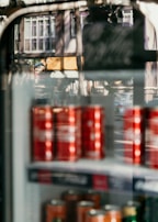 A refrigerator display filled with red cans is visible, with reflections of a street scene including buildings and cars on the glass. The photo combines both the indoor and outdoor environments, creating an interesting overlap of textures and colors.