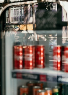 A refrigerator display filled with red cans is visible, with reflections of a street scene including buildings and cars on the glass. The photo combines both the indoor and outdoor environments, creating an interesting overlap of textures and colors.