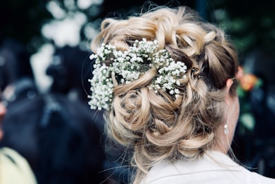 A view of an intricately styled updo featuring wavy, blonde hair intertwined with small white flowers. The arrangement appears elegant and suitable for a formal occasion.