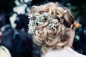 A view of an intricately styled updo featuring wavy, blonde hair intertwined with small white flowers. The arrangement appears elegant and suitable for a formal occasion.
