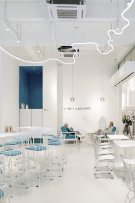 A modern and minimalist caf&eacute; with white walls and decorative lighting. The seating area includes white tables and chairs with blue upholstery. Two elderly people are sitting comfortably in blue chairs, engaged in a conversation. The space is bright and open, creating a serene atmosphere.