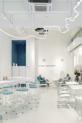 A modern and minimalist café with white walls and decorative lighting. The seating area includes white tables and chairs with blue upholstery. Two elderly people are sitting comfortably in blue chairs, engaged in a conversation. The space is bright and open, creating a serene atmosphere.