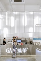 A modern and sleek cafe counter with a metallic finish dominates the foreground. Two employees, dressed in black uniforms, are busy preparing beverages. The counter is well-organized with a variety of equipment and supplies, including a coffee machine, cups, and product displays. Above the counter, there are bright, white lights that enhance the contemporary feel.