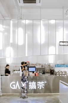 A modern and sleek cafe counter with a metallic finish dominates the foreground. Two employees, dressed in black uniforms, are busy preparing beverages. The counter is well-organized with a variety of equipment and supplies, including a coffee machine, cups, and product displays. Above the counter, there are bright, white lights that enhance the contemporary feel.