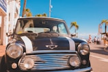 A classic black car with white racing stripes is parked on a sunny street near palm trees. The bright blue sky in the background adds to the summery atmosphere, while pedestrians walk by on the sidewalk. Nearby buildings and street signs indicate a coastal or tourist area.