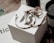 A set of jewelry items including large hoop earrings and a decorative ring are placed on top of a branded white box. The background features a dark-colored candle and some out-of-focus text, creating an elegant and sophisticated setting.