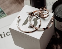 A set of jewelry items including large hoop earrings and a decorative ring are placed on top of a branded white box. The background features a dark-colored candle and some out-of-focus text, creating an elegant and sophisticated setting.