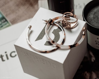 A set of jewelry items including large hoop earrings and a decorative ring are placed on top of a branded white box. The background features a dark-colored candle and some out-of-focus text, creating an elegant and sophisticated setting.