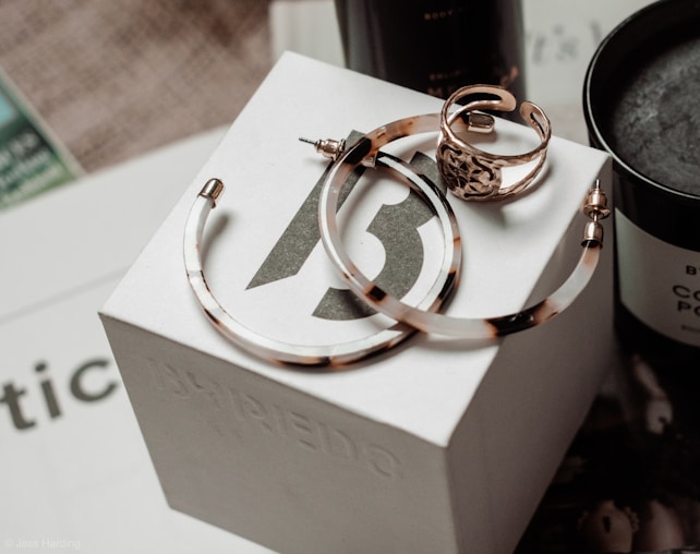 A set of jewelry items including large hoop earrings and a decorative ring are placed on top of a branded white box. The background features a dark-colored candle and some out-of-focus text, creating an elegant and sophisticated setting.