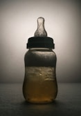 A baby bottle is depicted with textured liquid inside, illuminated from behind creating a silhouette. The background is soft and neutral, emphasizing the shape and details of the bottle.