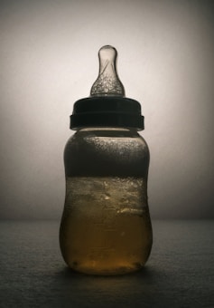 A baby bottle is depicted with textured liquid inside, illuminated from behind creating a silhouette. The background is soft and neutral, emphasizing the shape and details of the bottle.