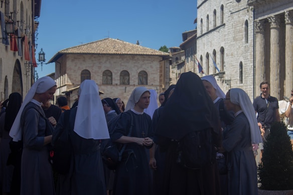 A group of nuns dressed in traditional habits gathers in a historical European town square. The background features old stone buildings with arched windows and columns, suggesting an ancient or classical architectural style. Other people are seen walking around, and the sunny day casts clear shadows.