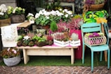 A display of various potted plants and flowers arranged creatively outside a store. The setup includes a wooden bench holding blooming plants in rectangular wooden crates, a small table draped with a lace cloth displaying more greenery, and a weathered turquoise chair with a white wire basket holding purple and green ornamental cabbages. The background includes large pots filled with plants and flowers, and the foreground features a basket of pink flowers. A sign on the chair indicates a price of five dollars.