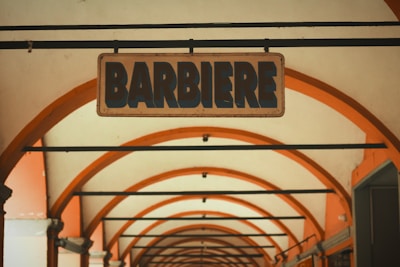 A vintage-style sign with the word 'Barbiere' hangs under a series of symmetrical archways. The arches are painted with warm orange and cream tones, creating a sense of depth and perspective.