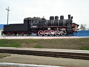 A black steam locomotive is prominently displayed on a raised platform beside railway tracks. The locomotive features red wheels with white rims, and there are white streaks accentuating its boiler. In the background, there is a blue and white building, along with some greenery.