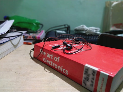 A red book titled 'The Art of Electronics' lies on a wooden desk with a pair of earphones placed on top. The background includes a green bottle, a stack of other books or files, and some electronics equipment.