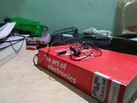 A red book titled 'The Art of Electronics' lies on a wooden desk with a pair of earphones placed on top. The background includes a green bottle, a stack of other books or files, and some electronics equipment.