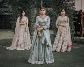 Three women are dressed in traditional Indian attire, standing in a grassy outdoor setting. The central woman wears a light blue lehenga with intricate embroidery, while the women on either side wear similarly ornate outfits in pastel shades. They are adorned with jewelry, including necklaces and earrings.