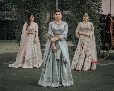 Three women are dressed in traditional Indian attire, standing in a grassy outdoor setting. The central woman wears a light blue lehenga with intricate embroidery, while the women on either side wear similarly ornate outfits in pastel shades. They are adorned with jewelry, including necklaces and earrings.