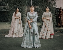 Three women are dressed in traditional Indian attire, standing in a grassy outdoor setting. The central woman wears a light blue lehenga with intricate embroidery, while the women on either side wear similarly ornate outfits in pastel shades. They are adorned with jewelry, including necklaces and earrings.