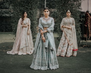 Three women are dressed in traditional Indian attire, standing in a grassy outdoor setting. The central woman wears a light blue lehenga with intricate embroidery, while the women on either side wear similarly ornate outfits in pastel shades. They are adorned with jewelry, including necklaces and earrings.