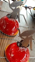 Two glossy, red dome-shaped desserts rest on a cooling rack. Each is topped with intricate chocolate sculptures resembling abstract art. Small sugar crystals are sprinkled at the base of the chocolate pieces, adding a touch of elegance.
