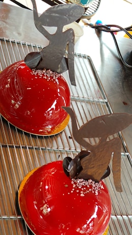 Two glossy, red dome-shaped desserts rest on a cooling rack. Each is topped with intricate chocolate sculptures resembling abstract art. Small sugar crystals are sprinkled at the base of the chocolate pieces, adding a touch of elegance.