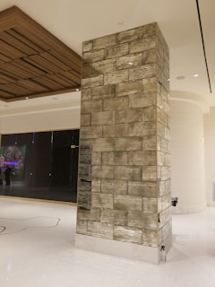 A modern interior space featuring a tall rectangular column covered in a textured tile pattern. The column is situated on a glossy white floor, and in the background, there is a large glass panel with a colorful display and overhead wooden ceiling detail.