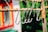 Several metallic S-hooks are hanging on a rusted horizontal rod. The background features a blurred mix of green foliage and part of a colorful building, possibly red and yellow. The focus is on the hooks, emphasizing their shiny surface against the rustic rod.