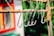 Several metallic S-hooks are hanging on a rusted horizontal rod. The background features a blurred mix of green foliage and part of a colorful building, possibly red and yellow. The focus is on the hooks, emphasizing their shiny surface against the rustic rod.