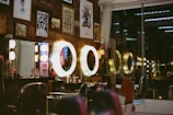 A vintage-style barbershop interior is adorned with framed artwork and illuminated by warm, circular lights. Several barber chairs are positioned in front of the mirrors, and various grooming products are visible on the countertops. The wooden wall and the reflection of neon signs add to the retro ambiance of the space.