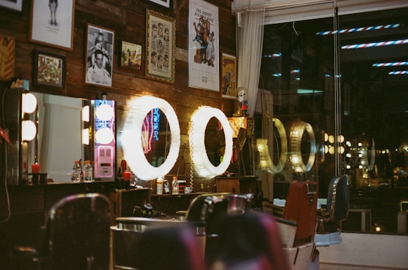 A vintage-style barbershop interior is adorned with framed artwork and illuminated by warm, circular lights. Several barber chairs are positioned in front of the mirrors, and various grooming products are visible on the countertops. The wooden wall and the reflection of neon signs add to the retro ambiance of the space.