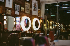 A vintage-style barbershop interior is adorned with framed artwork and illuminated by warm, circular lights. Several barber chairs are positioned in front of the mirrors, and various grooming products are visible on the countertops. The wooden wall and the reflection of neon signs add to the retro ambiance of the space.
