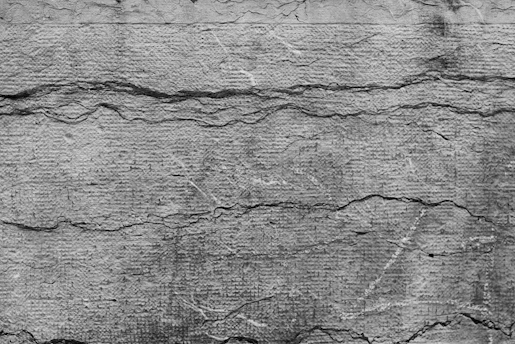 Rough textured concrete wall with visible cracks running horizontally and vertically. The surface appears weathered and aged, with various shades of gray contributing to an overall rustic and rugged look.