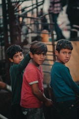 Three young boys stand together, each wearing casual clothing. The atmosphere appears earthy and muted, possibly due to the surrounding industrial or urban environment. The boys are looking towards the camera with neutral expressions.