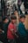 Three young boys stand together, each wearing casual clothing. The atmosphere appears earthy and muted, possibly due to the surrounding industrial or urban environment. The boys are looking towards the camera with neutral expressions.