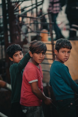 Three young boys stand together, each wearing casual clothing. The atmosphere appears earthy and muted, possibly due to the surrounding industrial or urban environment. The boys are looking towards the camera with neutral expressions.