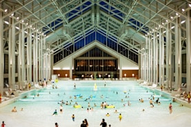A spacious indoor pool area featuring a high, glass-paneled ceiling and well-lit surroundings. Numerous people of various ages are swimming, floating, or relaxing in the large swimming pool. The architectural design is modern and open, with tall supporting beams and a symmetrical layout. There are pool chairs lining the sides and a few decorations, such as potted plants, near the back wall.