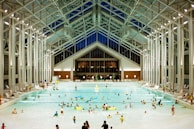 A spacious indoor pool area featuring a high, glass-paneled ceiling and well-lit surroundings. Numerous people of various ages are swimming, floating, or relaxing in the large swimming pool. The architectural design is modern and open, with tall supporting beams and a symmetrical layout. There are pool chairs lining the sides and a few decorations, such as potted plants, near the back wall.