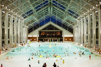 A spacious indoor pool area featuring a high, glass-paneled ceiling and well-lit surroundings. Numerous people of various ages are swimming, floating, or relaxing in the large swimming pool. The architectural design is modern and open, with tall supporting beams and a symmetrical layout. There are pool chairs lining the sides and a few decorations, such as potted plants, near the back wall.