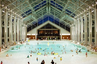 A spacious indoor pool area featuring a high, glass-paneled ceiling and well-lit surroundings. Numerous people of various ages are swimming, floating, or relaxing in the large swimming pool. The architectural design is modern and open, with tall supporting beams and a symmetrical layout. There are pool chairs lining the sides and a few decorations, such as potted plants, near the back wall.