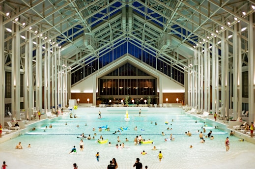 A spacious indoor pool area featuring a high, glass-paneled ceiling and well-lit surroundings. Numerous people of various ages are swimming, floating, or relaxing in the large swimming pool. The architectural design is modern and open, with tall supporting beams and a symmetrical layout. There are pool chairs lining the sides and a few decorations, such as potted plants, near the back wall.