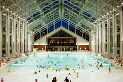A spacious indoor pool area featuring a high, glass-paneled ceiling and well-lit surroundings. Numerous people of various ages are swimming, floating, or relaxing in the large swimming pool. The architectural design is modern and open, with tall supporting beams and a symmetrical layout. There are pool chairs lining the sides and a few decorations, such as potted plants, near the back wall.