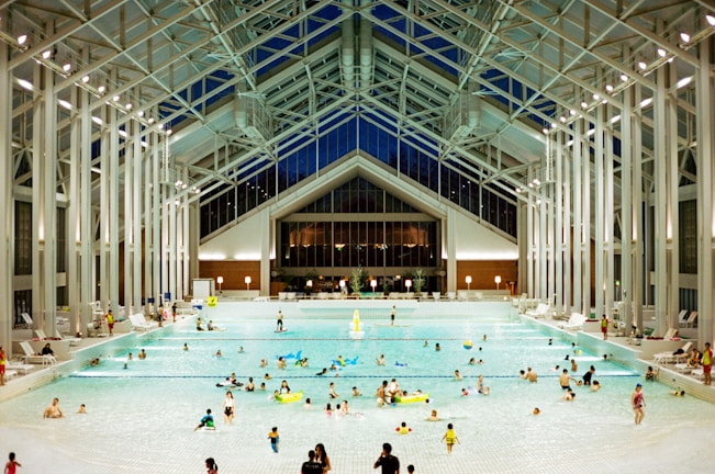 A spacious indoor pool area featuring a high, glass-paneled ceiling and well-lit surroundings. Numerous people of various ages are swimming, floating, or relaxing in the large swimming pool. The architectural design is modern and open, with tall supporting beams and a symmetrical layout. There are pool chairs lining the sides and a few decorations, such as potted plants, near the back wall.