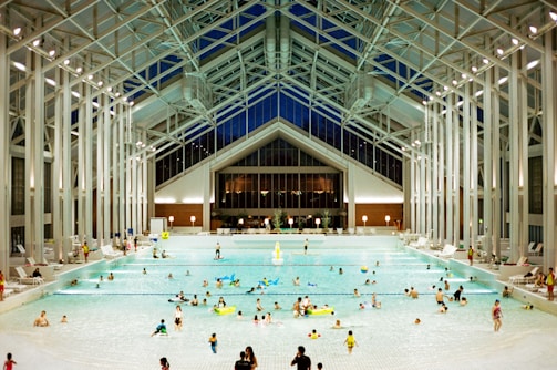 A spacious indoor pool area featuring a high, glass-paneled ceiling and well-lit surroundings. Numerous people of various ages are swimming, floating, or relaxing in the large swimming pool. The architectural design is modern and open, with tall supporting beams and a symmetrical layout. There are pool chairs lining the sides and a few decorations, such as potted plants, near the back wall.